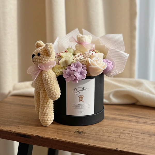 Floral arrangement in a black box with a small crocheted bear on a wooden table.